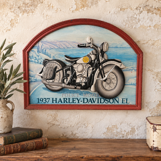 A framed wall art piece featuring a vintage 1937 Harley-Davidson motorcycle illustration, displayed on a rustic wall above a wooden shelf with books and a plant.