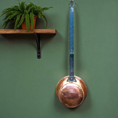 Copper ladle hanging on a green wall with a wooden shelf and plant.