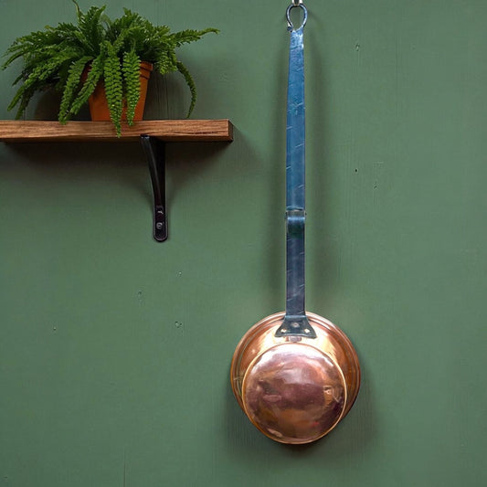 Copper ladle hanging on a green wall with a wooden shelf and plant.