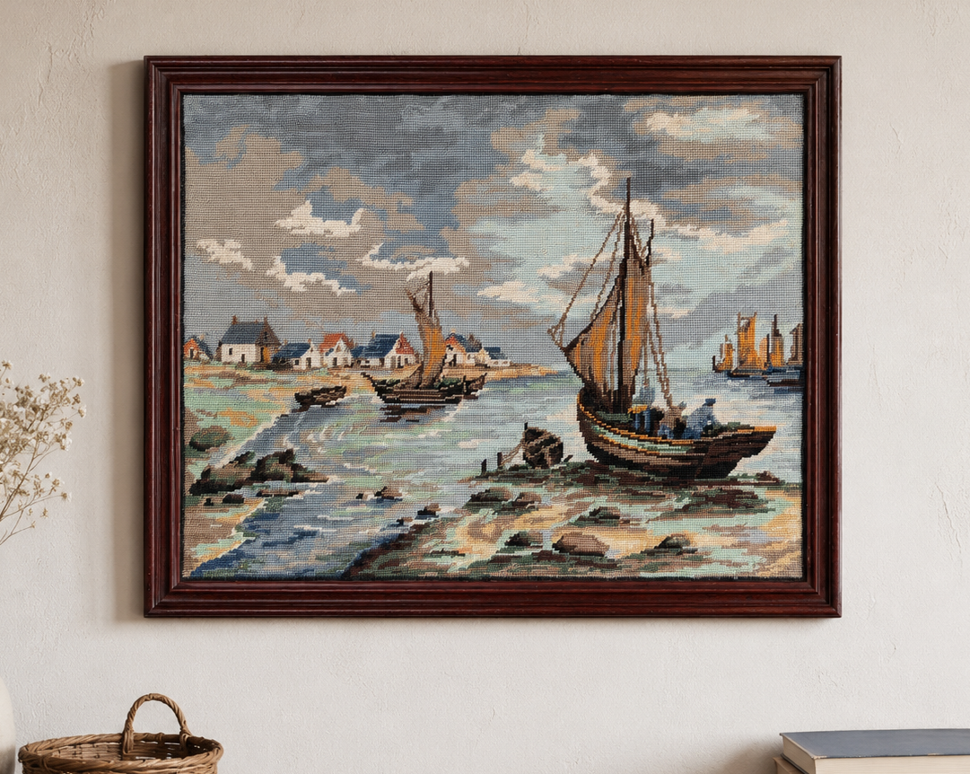 Framed tapestry needlepoint of boats on a beach with a vase and books on a wooden shelf below.