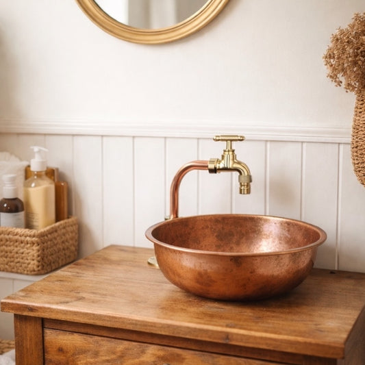 Copper sink with gold faucet on a wooden cabinet against a white paneled wall.