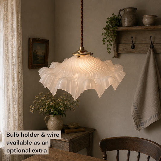 A vintage-style pendant light with a ruffled, translucent white glass shade and a brass-toned cap, hanging in a cozy, rustic dining room setting. A text overlay in the bottom-left corner reads 'Bulb holder & wire available as an optional extra'.