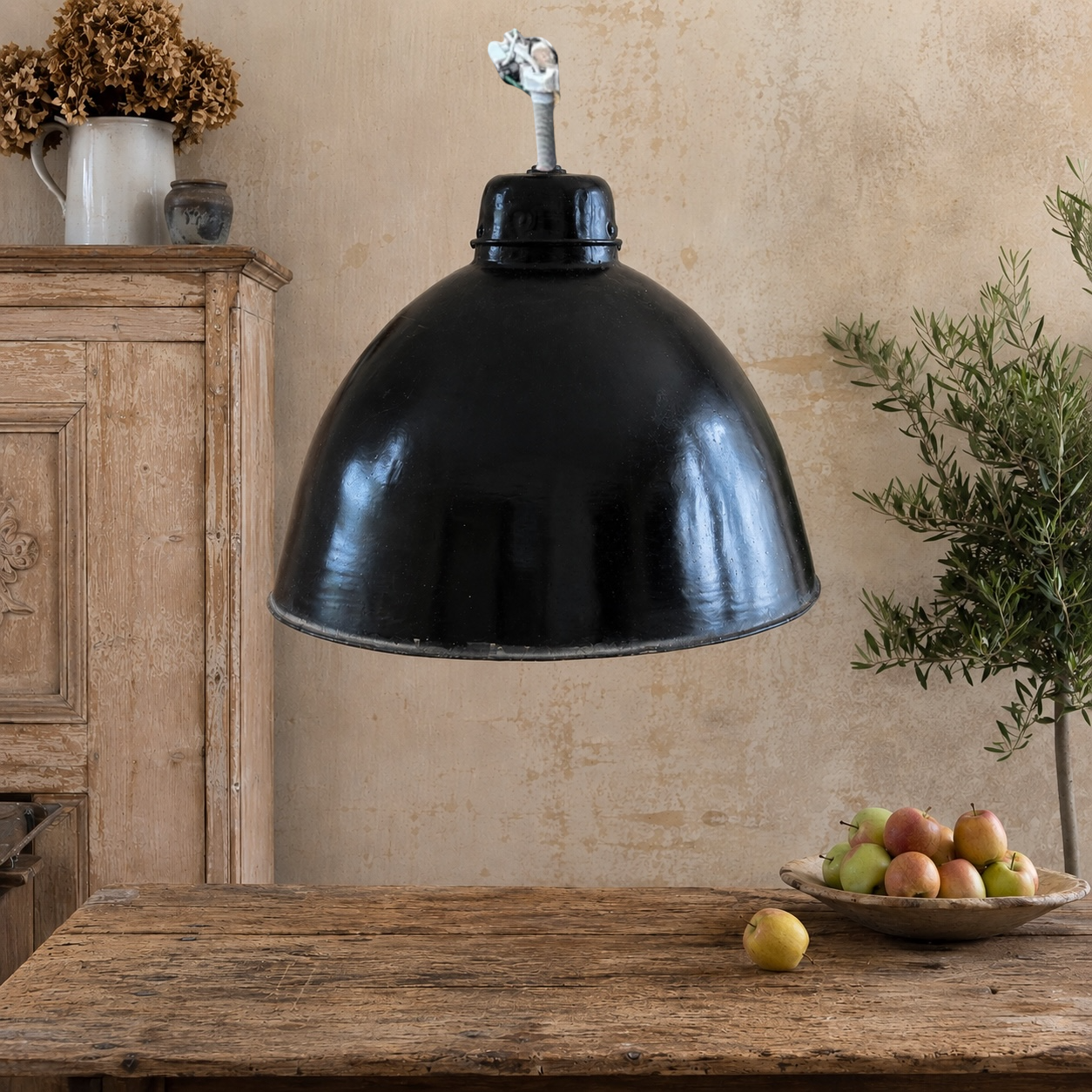 A large black metal dome pendant light hanging over a rustic wooden table in a styled room with a textured beige wall, a wooden cabinet, and a bowl of apples.