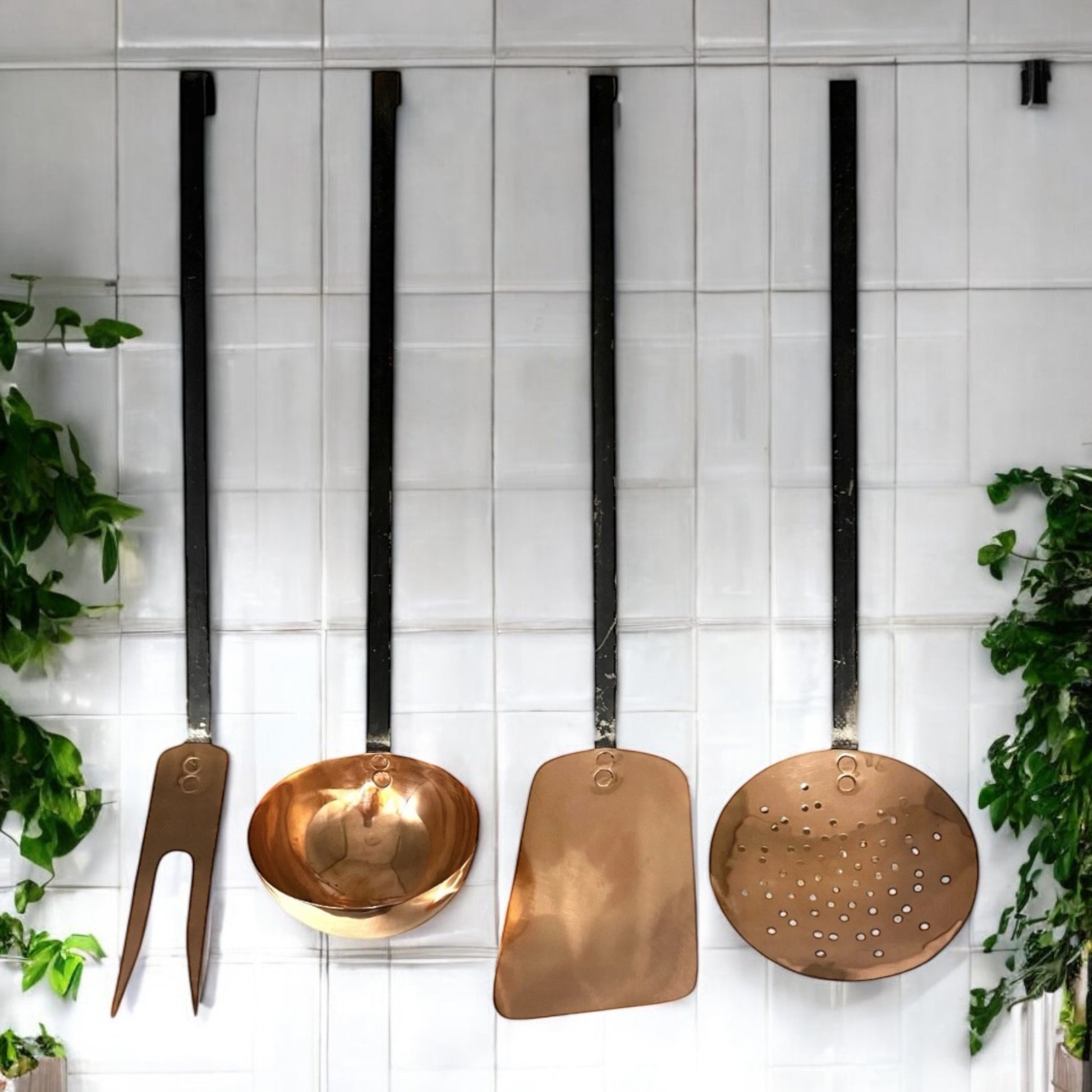 set of 4 French copper utensils with black steel handles against a white tiled wall with plants