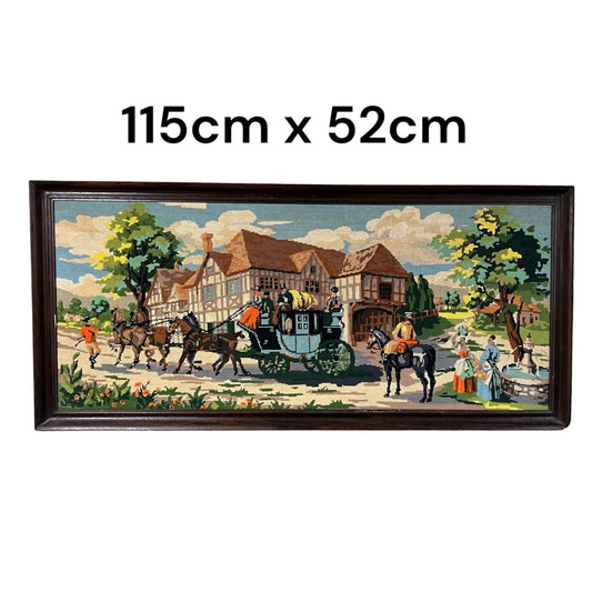 A framed needlepoint canvas featuring a village scene with a horse-drawn carriage, trees, and people, shown on a white background with the text '115cm x 52cm' above it.