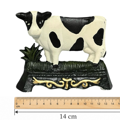 A black and white cow figurine standing on a dark green decorative base with a gold scroll motif, shown against a white background with a wooden ruler at the bottom indicating 14 cm width.