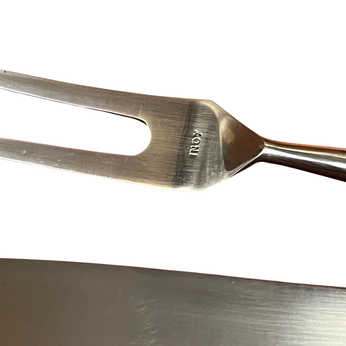 Close-up of a stainless steel carving fork with the brand name 'INOX' engraved on the blade, set against a white background with a dark metallic surface at the bottom.