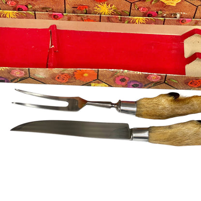 A two-piece serving utensil set featuring a stainless steel carving fork and knife with natural animal hide handles, displayed alongside a red velvet-lined box with a floral-patterned lid on a white background.