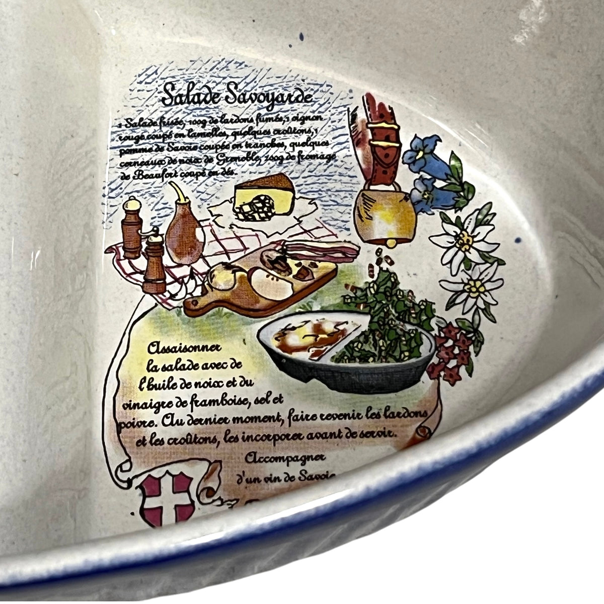 Close-up of the inside of a ceramic bowl or dish featuring a printed French recipe illustration for Salade Savoyarde, with illustrated ingredients, text, and a blue rim visible along the edge.