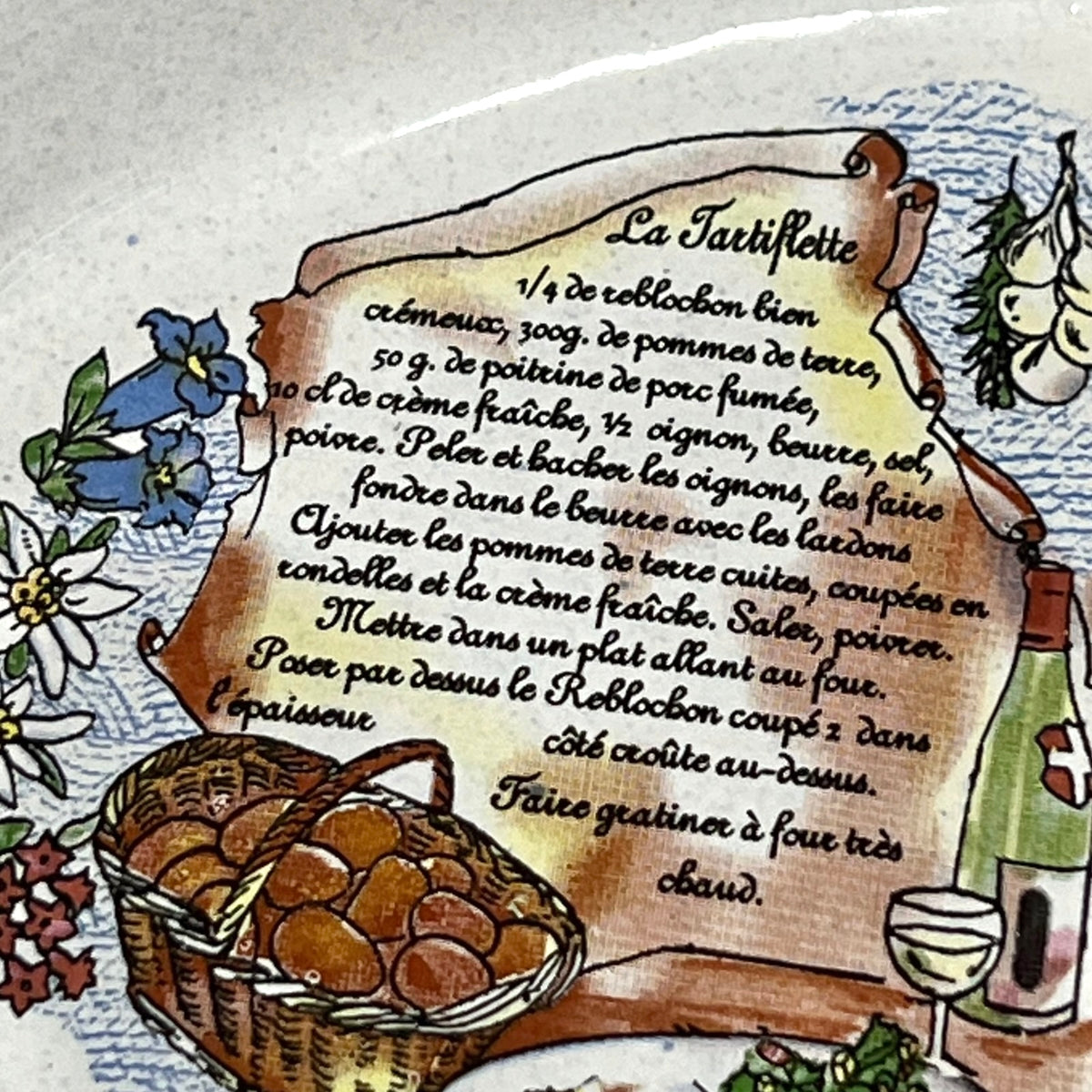 A close-up of a decorative ceramic plate or dish featuring a hand-painted recipe for 'La Tartiflette' on a parchment scroll, surrounded by illustrations of eggs, flowers, and a wine glass.