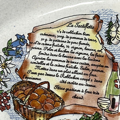 A close-up of a decorative ceramic plate or dish featuring a hand-painted recipe for 'La Tartiflette' on a parchment scroll, surrounded by illustrations of eggs, flowers, and a wine glass.