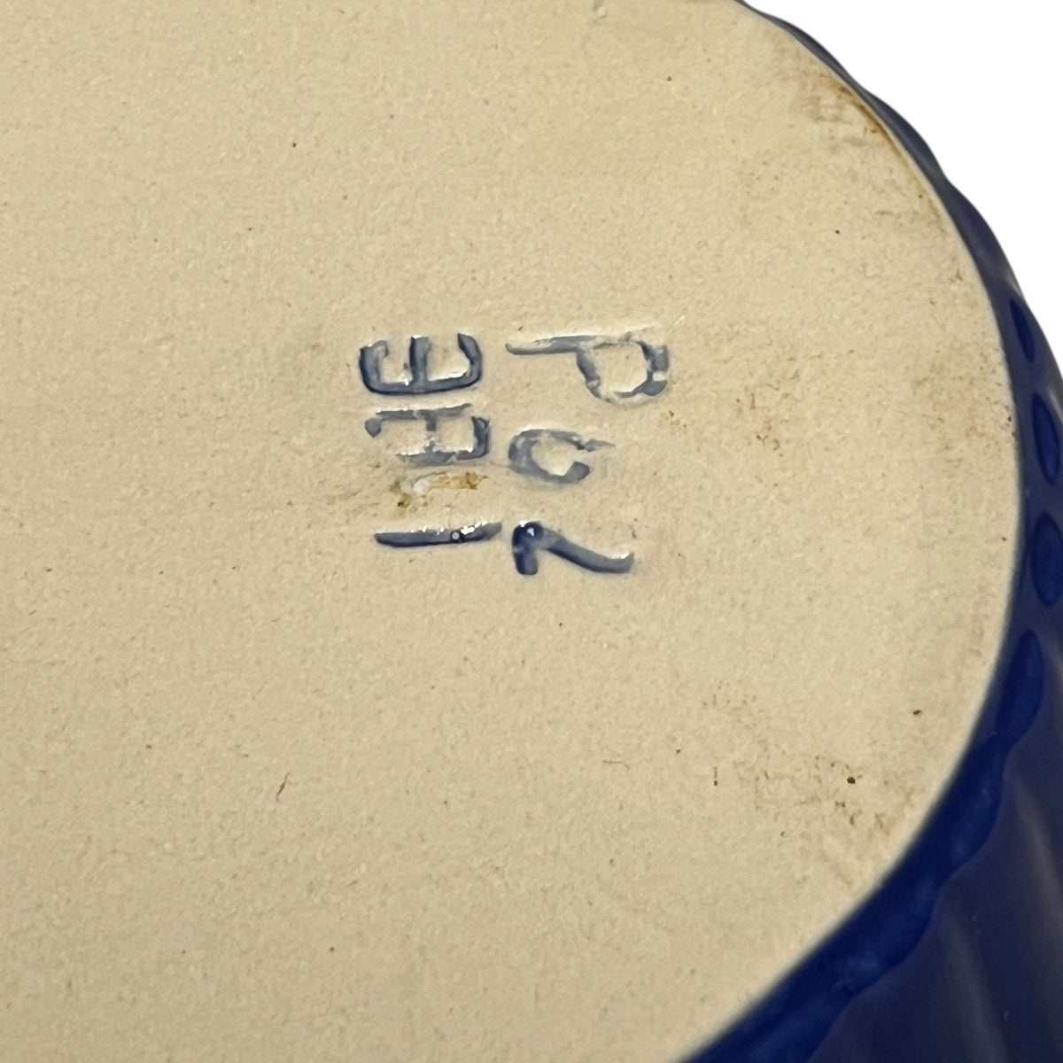Close-up of the bottom of a ceramic dish showing a blue hand-stamped maker's mark on a beige glaze, with a dark blue rim visible on the right.