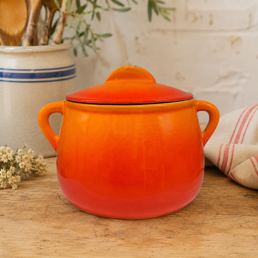 An orange ceramic Le Creuset lidded pot with two side handles sits on a wooden tabletop, with a white brick wall, kitchen utensils, and a striped cloth in the background.