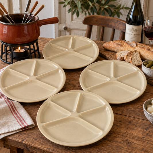 Four round ceramic divided serving plates in a warm cream-beige color, arranged on a rustic wooden dining table with bread, a wine bottle, a fondue pot, and small bowls in a styled indoor setting.