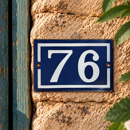 A rectangular blue house number plaque with white lettering reading '76', mounted on a textured beige stone wall next to a weathered blue wooden door and green leaves.