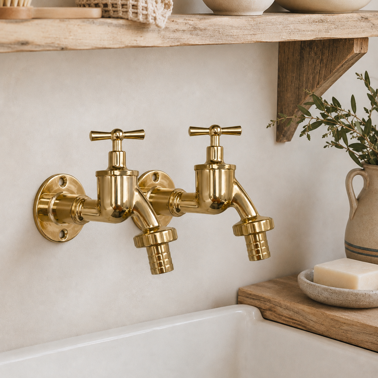 Two brass wall-mounted tap faucets with cross handles, installed on a beige stucco wall above a white sink, with a wooden shelf and decorative ceramic items in the background.