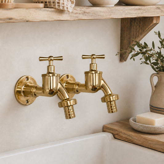 Two brass wall-mounted tap faucets with cross handles, installed on a beige stucco wall above a white sink, with a wooden shelf and decorative ceramic items in the background.