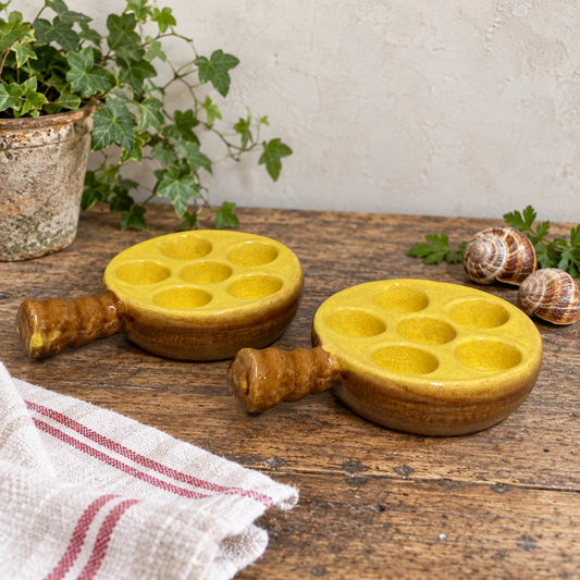 Pair of vintage French ceramic escargot dishes in warm mustard yellow and rustic brown glazed stoneware, each featuring six recessed wells for serving traditional French snails and a short integrated handle. The dishes measure 12cm in diameter, 16cm including the handle and 3.5cm deep. Displayed on a rustic wooden kitchen table with a trailing ivy plant, traditional French striped linen tea towel and two edible snail shells with parsley. A charming French brocante find