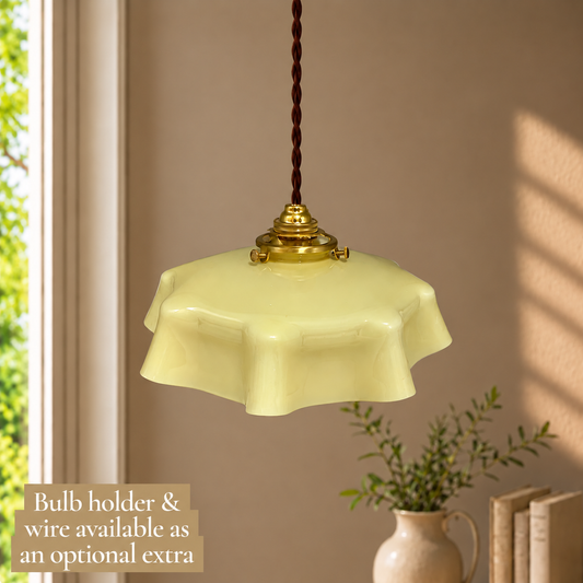 A vintage-style pendant light with a pale yellow glass shade and a brass-colored metal fixture, hanging from a dark brown cord in a warm, sunlit room. A text overlay in the bottom-left corner reads 'Bulb holder & wire available as an optional extra'.