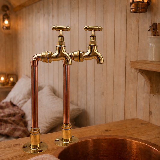 A rustic brass and copper sink faucet set installed above a copper vessel sink on a wooden countertop inside a cozy wood-paneled room with warm ambient lighting.