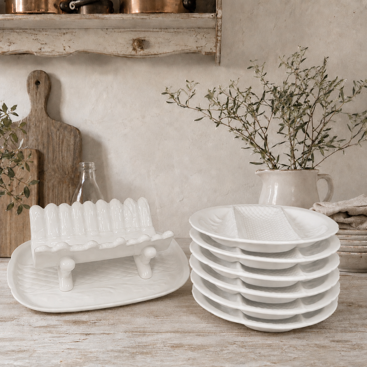 Stacked white ceramic divided serving plates and a matching white ceramic asparagus serving dish displayed on a rustic wooden table in a French farmhouse-style kitchen. The scene features weathered wooden boards, olive branches in a cream jug, and vintage copper cookware, creating a warm and elegant country kitchen setting.