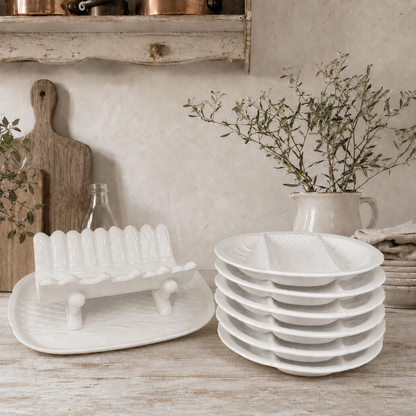 Stacked white ceramic divided serving plates and a matching white ceramic asparagus serving dish displayed on a rustic wooden table in a French farmhouse-style kitchen. The scene features weathered wooden boards, olive branches in a cream jug, and vintage copper cookware, creating a warm and elegant country kitchen setting.