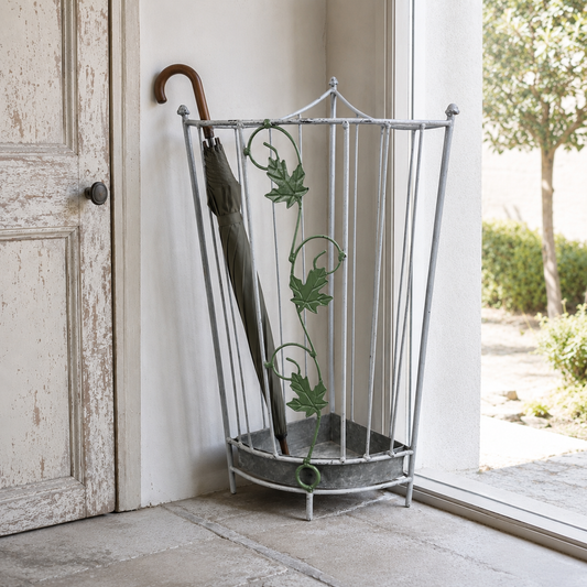 A tall metal umbrella stand with decorative green vine and leaf motifs, holding a dark umbrella, positioned next to a distressed white door in a doorway.