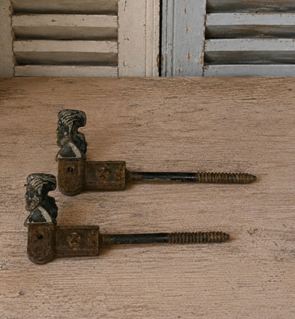 A pair of vintage metal shutter dogs with sculpted busts at the top and threaded screw backs, displayed on a rustic wooden surface.