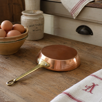 Small French copper egg pan with a brass handle on a farmhouse kitchen table 