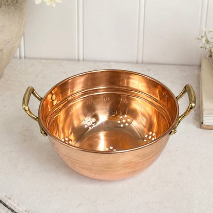 A copper colander with two brass handles, shown on a light stone countertop with a white panelled backsplash in the background.