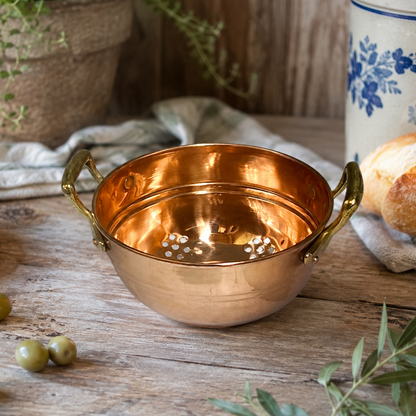 A copper colander with two handles sits on a rustic wooden table, surrounded by olives, bread, a potted plant, and a ceramic container with blue floral detailing.