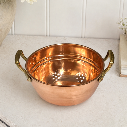 A copper with two brass handles, shown on a light stone countertop against a white paneled wall.