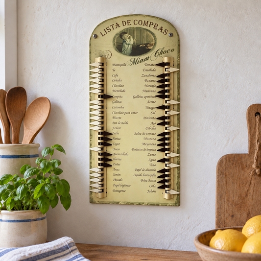 A vintage-style Spanish 'Lista de Compras' shopping list wall organizer hanging on a textured white wall in a kitchen setting. The organizer features a distressed beige background with black and white  tabs, and the name 'Miam Choco' written at the top.