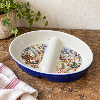 A ceramic divided serving dish with a deep blue ribbed base and a white interior featuring whimsical food illustrations and recipe text, shown on a wooden tabletop with a plant and striped cloth nearby.