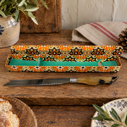 A decorative bread knife with a patterned box set on a rustic wooden table. The box features a vibrant retro floral pattern in orange, black, white, and green, with a bright green interior.