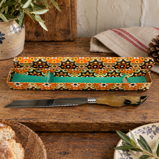 A decorative bread knife with a patterned box set on a rustic wooden table. The box features a vibrant retro floral pattern in orange, black, white, and green, with a bright green interior.