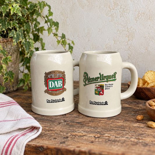 Two ceramic beer steins on a rustic wooden table, one with a DAB logo and the other with a Piner Urquell logo, surrounded by a plant, chips, and a striped cloth.