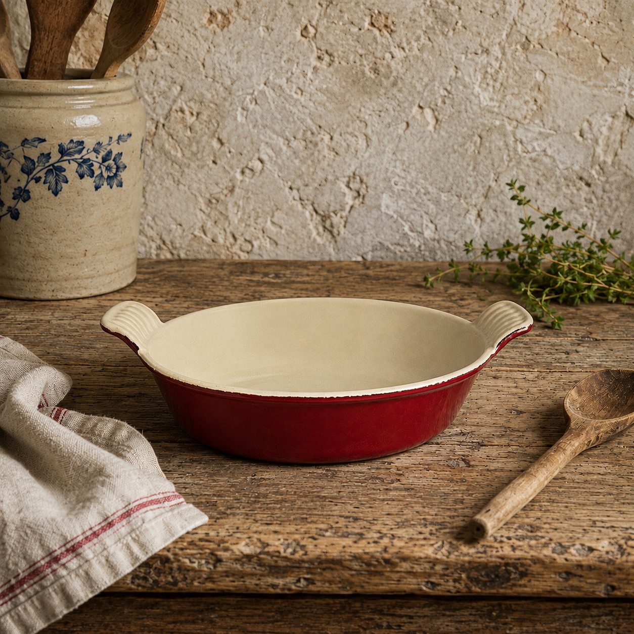 A red and cream enameled baking dish with side handles sits on a rustic wooden table, surrounded by a linen napkin, wooden spoon, ceramic utensil jar, and a small sprig of herbs against a textured plaster wall.