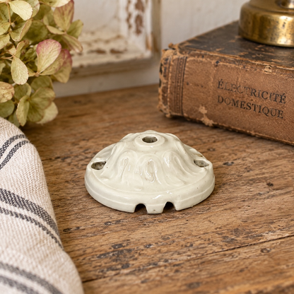 A small off-white ceramic decorative ceiling rose with three screw holes sits on a rustic wooden table, styled with a vintage book, a striped cloth, and dried flowers.