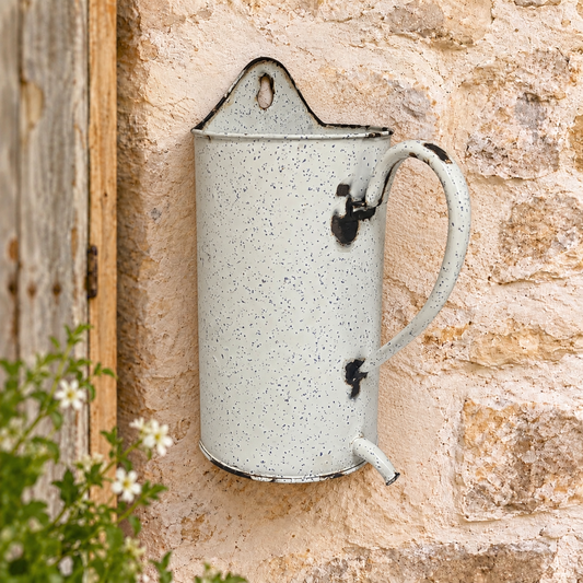 A vintage-style speckled enamel planter mounted on a rustic stone wall, with a wooden door and small white flowers visible at the side.