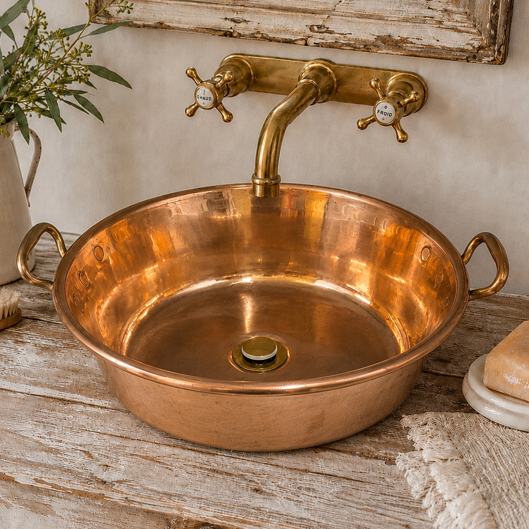 A round copper vessel sink with side handles and a matching brass faucet, set on a distressed wooden vanity with decorative props.