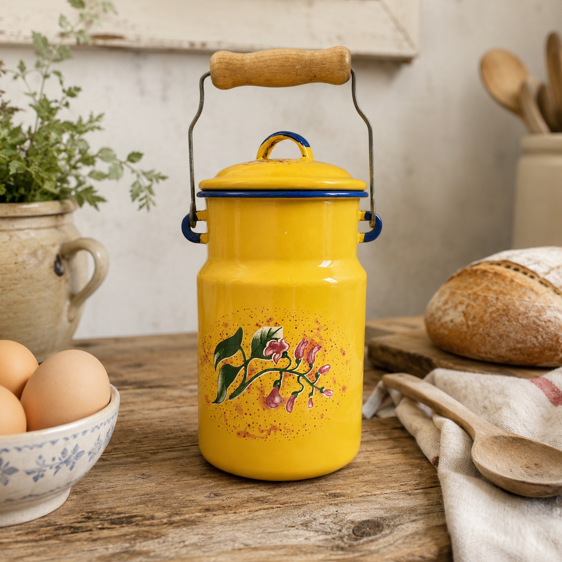 A bright yellow enamel coffee kettle with a wooden handle and lid, decorated with a pink and green floral design, set on a rustic wooden table surrounded by bread, eggs, and kitchen items.