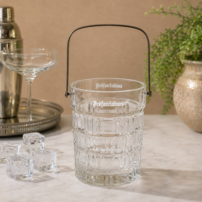 A clear glass ice bucket with a black metal handle, featuring the brand name 'Préfontaines' printed on the front, set on a marble countertop with ice cubes, a cocktail shaker, and a coupe glass in a warm, styled setting.
