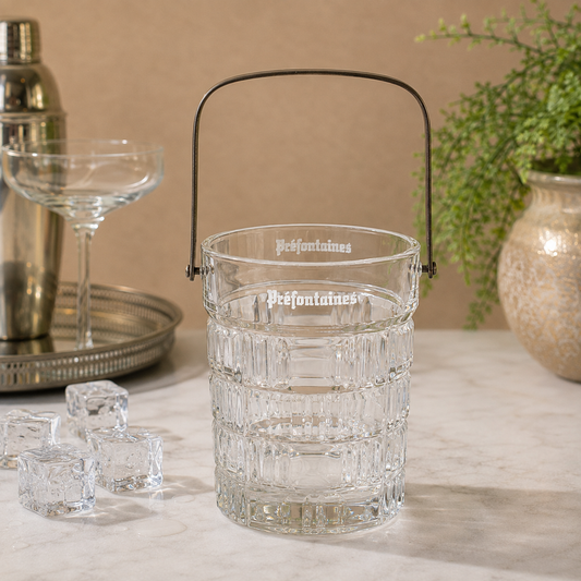 A clear glass ice bucket with a black metal handle, featuring the brand name 'Préfontaines' printed on the front, set on a marble countertop with ice cubes, a cocktail shaker, and a coupe glass in a warm, styled setting.