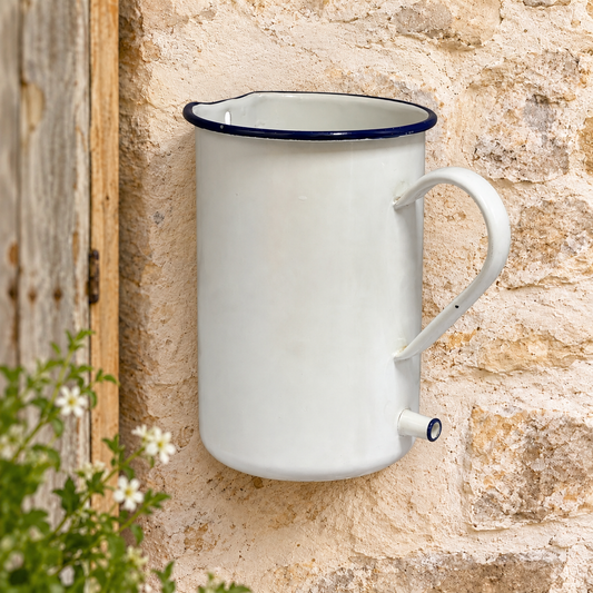 A vintage-style white enamel wall-mounted planter with a dark blue rim and spout, mounted on a rustic beige stone wall next to a wooden door.