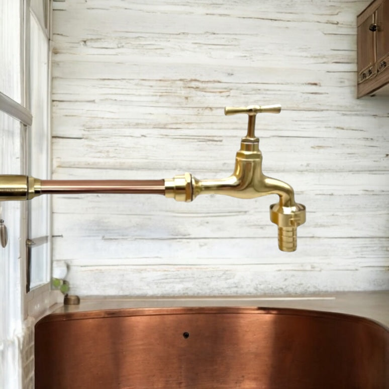 A brass wall-mounted faucet with a cross handle and copper pipe, installed above a copper sink against a white rustic wood plank wall.