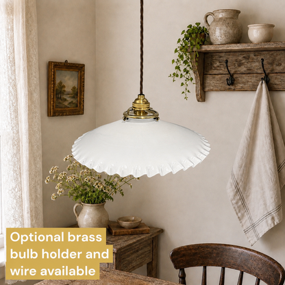 A white ruffled glass pendant light hanging in a rustic dining room setting with a wooden chair, side table, and wall decor. A text overlay in the bottom left corner reads 'Optional brass bulb holder and wire available'.