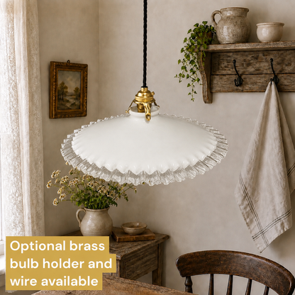 A white ruffled glass pendant light hanging in a rustic dining room setting with a wooden chair, side table, and wall decor. A text overlay in the bottom left corner reads 'Optional brass bulb holder and wire available'.
