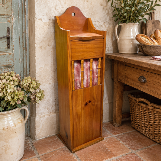 A tall wooden baguette storage box with a slatted front and red-and-white gingham fabric panel, shown in a rustic kitchen setting next to a wooden table and tiled floor.