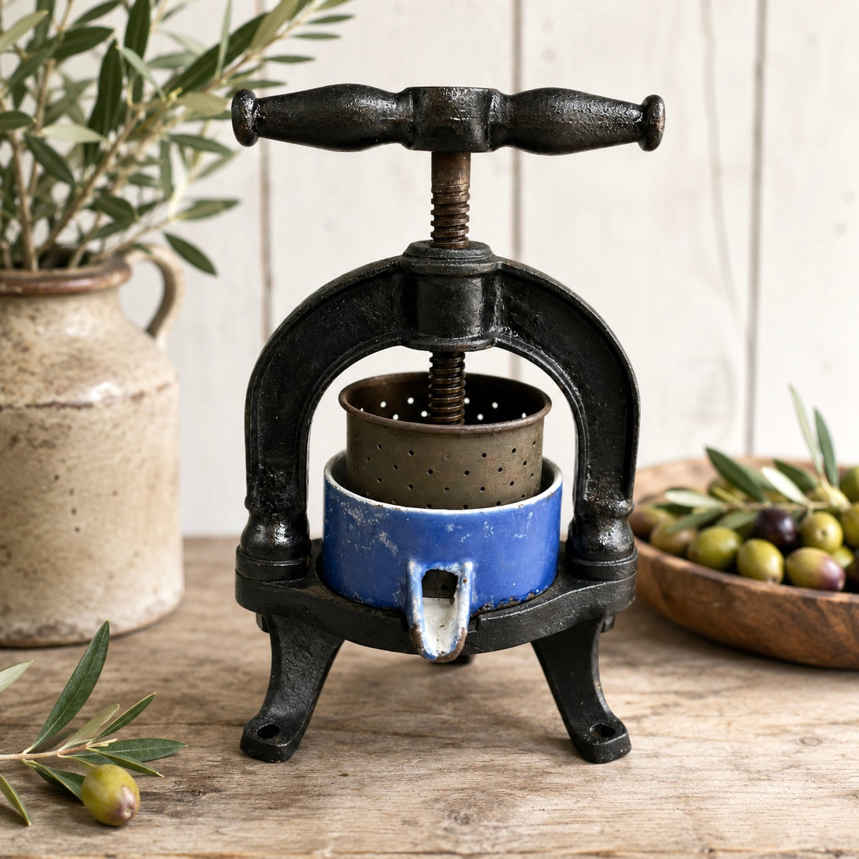 Vintage-style olive press on a wooden surface with olives and a plant in the background.